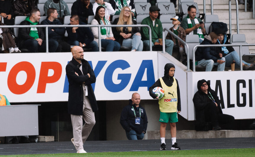 Coach with arms crossed on the sideline as a youth player in a yellow bib holds a soccer ball nearby; crowd in the stands behind them.