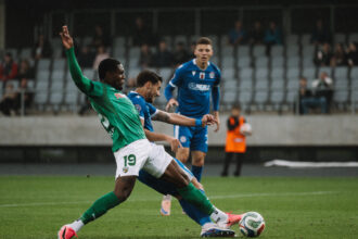 Soccer players in green and blue jerseys battle for the ball on a grass field with spectators in the stands behind them.