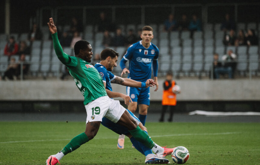 Soccer players in green and blue jerseys battle for the ball on a grass field with spectators in the stands behind them.