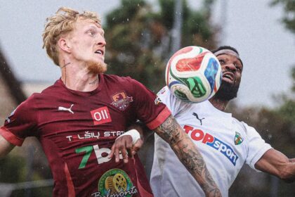 Two soccer players vie for a header as the ball approaches, in a rainy outdoor match, both reaching for it.