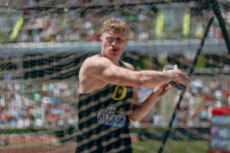Male track-and-field athlete in a throwing cage, mid-throw with a discus, wearing a black singlet and bib in bright sunlight.