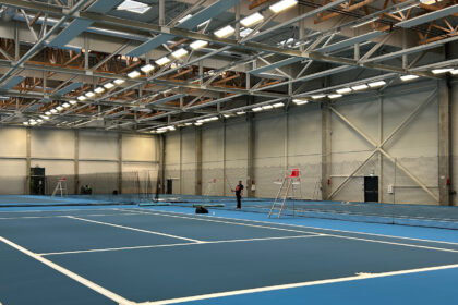 Indoor tennis facility with blue courts, white court lines, and a person standing near the net under bright overhead lights, showing a large training hall.