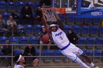 Basketball player in a white jersey dunks the ball over the rim as spectators watch from blue-seated arena.