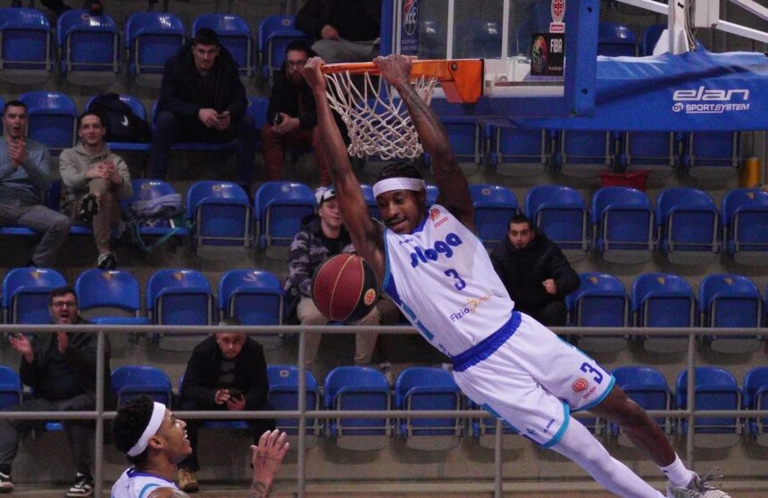 Basketball player in a white jersey dunks the ball over the rim as spectators watch from blue-seated arena.