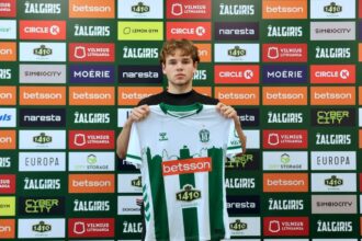 Young male soccer player holding up a green-and-white striped jersey in front of a sponsor-filled backdrop, announcing team affiliation.