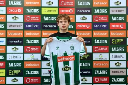 Young male soccer player holding up a green-and-white striped jersey in front of a sponsor-filled backdrop, announcing team affiliation.