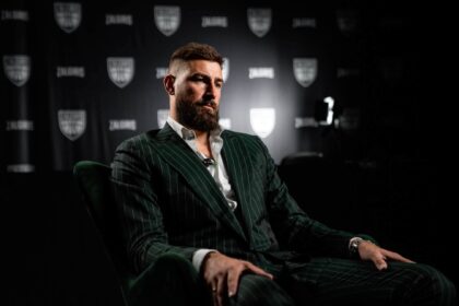 Man with a full beard in a dark pinstripe suit sits in a studio during an interview, backdrop featuring shield logos behind him.