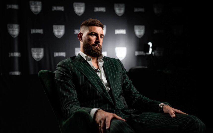 Man with a full beard in a dark pinstripe suit sits in a studio during an interview, backdrop featuring shield logos behind him.