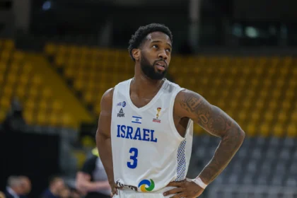 Basketball player in a white Israel jersey with number 3, hands on hips, standing on a court with yellow seats blurred in the background.