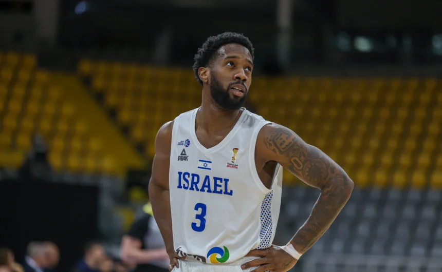 Basketball player in a white Israel jersey with number 3, hands on hips, standing on a court with yellow seats blurred in the background.