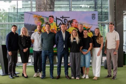 Group of ten people posing for a photo in front of a large sports banner indoors with a green carpeted floor in a modern space around them.
