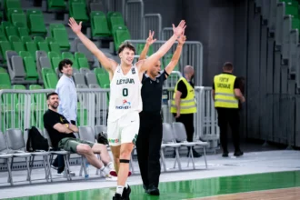 Lithuanian basketball player in a white jersey raises both arms in celebration on the sideline during a game.