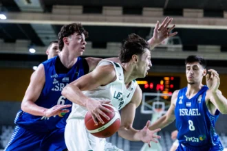 Basketball player in white drives with ball while defenders in blue Israel jerseys converge in an indoor court.