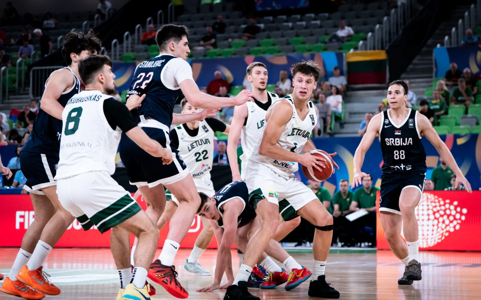 Basketball players fight for the ball on a crowded indoor court, scrambling near the baseline with fans in the stands behind them.