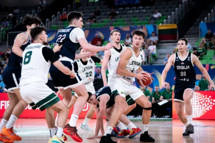 Basketball players fight for the ball on a crowded indoor court, scrambling near the baseline with fans in the stands behind them.
