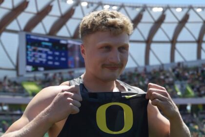 Sprinter on a track holding his black bib with a large yellow Oregon 'O' logo, with a stadium and scoreboard in the background.