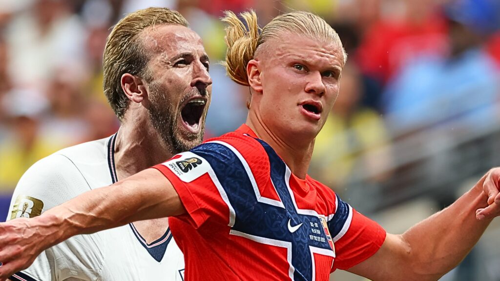 Two soccer players celebrate together on the field, one in red and blue and the other in white, arms raised in triumph against a blurred crowd background.