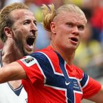 Two soccer players celebrate together on the field, one in red and blue and the other in white, arms raised in triumph against a blurred crowd background.