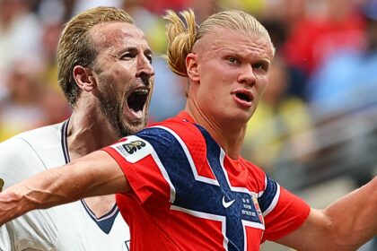 Two soccer players celebrate together on the field, one in red and blue and the other in white, arms raised in triumph against a blurred crowd background.
