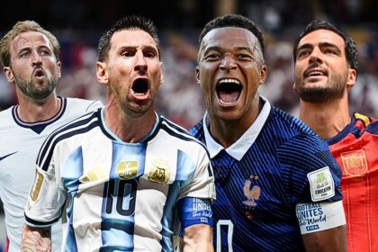 Four football players in national team jerseys cheer with mouths open, celebrating on the pitch with a stadium crowd behind them.