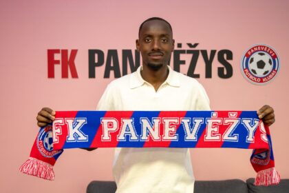 Man wearing a white polo holds a red and blue FK Panevėžys scarf in front of a pink wall with the club logo visible to the right.