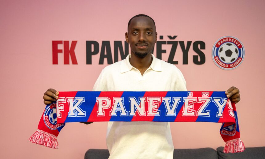 Man wearing a white polo holds a red and blue FK Panevėžys scarf in front of a pink wall with the club logo visible to the right.