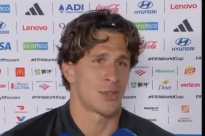 Male soccer player speaks to a mic during a post-match interview, backdrop filled with sponsor logos behind him.