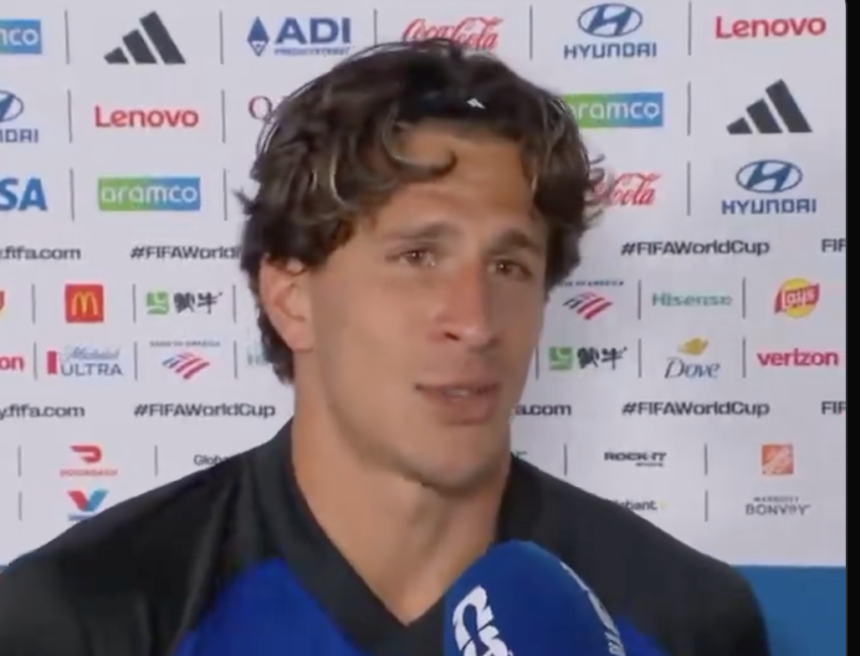 Male soccer player speaks to a mic during a post-match interview, backdrop filled with sponsor logos behind him.
