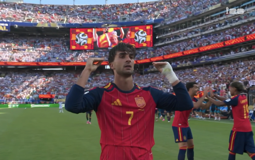 Spain player in red kit with number 7 celebrates on the pitch as fans cheer in stadium stands behind him.