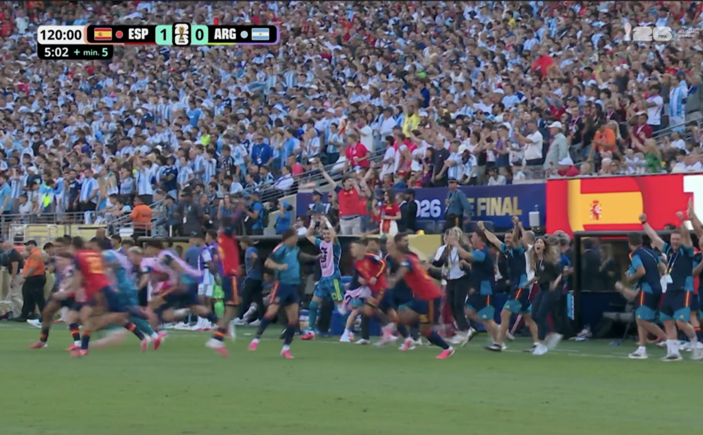 Crowd of spectators in a packed stadium watching a sprint on the field, scoreboard shows ESP 1 ARG 0 with a World Cup logo nearby.