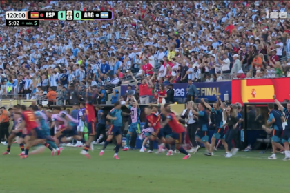Crowd of spectators in a packed stadium watching a sprint on the field, scoreboard shows ESP 1 ARG 0 with a World Cup logo nearby.