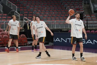 German basketball players in white jerseys on an indoor court; one player is holding a basketball while others walk nearby, empty red and gray seats in the background.