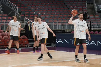 German basketball players in white jerseys on an indoor court; one player is holding a basketball while others walk nearby, empty red and gray seats in the background.
