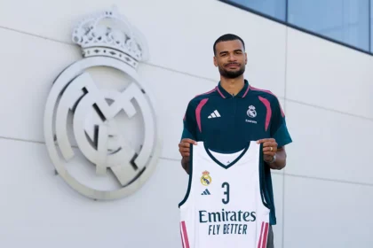 Male footballer stands outside a Real Madrid facility, holding a white Real Madrid jersey with number 3 and Emirates Fly Better in front of the club crest.