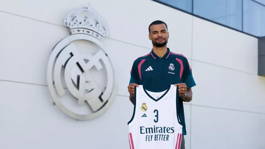 Male footballer stands outside a Real Madrid facility, holding a white Real Madrid jersey with number 3 and Emirates Fly Better in front of the club crest.