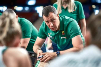 Man in a green polo shirt leans over a wooden table, writing with a marker while teammates watch at a sports event.