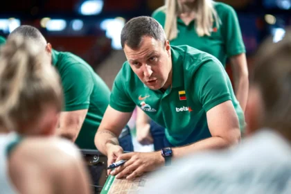 Man in a green polo shirt leans over a wooden table, writing with a marker while teammates watch at a sports event.
