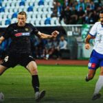 Two soccer players chase the ball during a match on a green field, one in a black kit and the other in white and blue.
