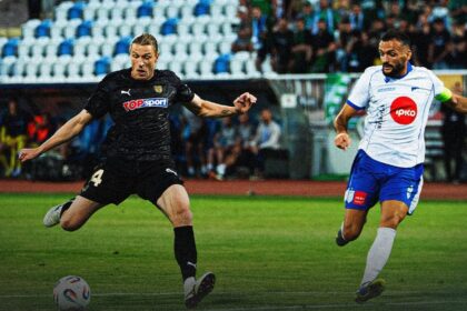 Two soccer players chase the ball during a match on a green field, one in a black kit and the other in white and blue.