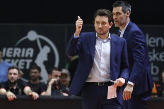 Two coaches in blue suits on a gym floor, one giving a thumbs-up and holding a pink note, with spectators watching behind them.