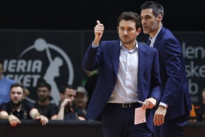 Two coaches in blue suits on a gym floor, one giving a thumbs-up and holding a pink note, with spectators watching behind them.