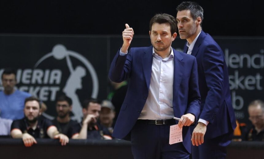 Two coaches in blue suits on a gym floor, one giving a thumbs-up and holding a pink note, with spectators watching behind them.