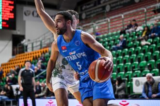 Male basketball player in a blue jersey drives with the ball during a game, stands filled with green seats in the background.