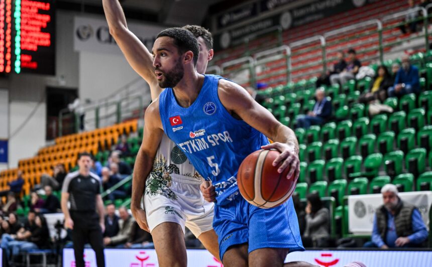 Male basketball player in a blue jersey drives with the ball during a game, stands filled with green seats in the background.