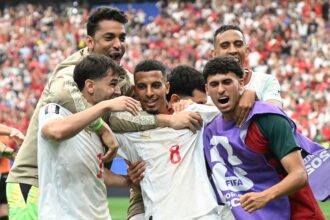 A group of soccer players in white and purple jerseys celebrate together on the field with a cheering crowd behind them.