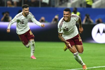 Two soccer players in white uniforms run and celebrate on a green pitch, one smiling as they sprint forward.