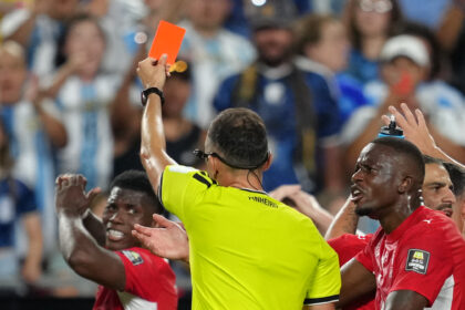 Soccer referee in a neon yellow shirt holds up an orange card to players as a crowd looks on behind him.
