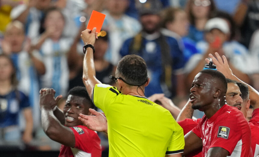 Soccer referee in a neon yellow shirt holds up an orange card to players as a crowd looks on behind him.