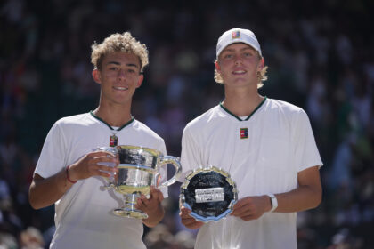 Two young athletes in white shirts pose with large trophies at a ceremony, smiling for the camera.