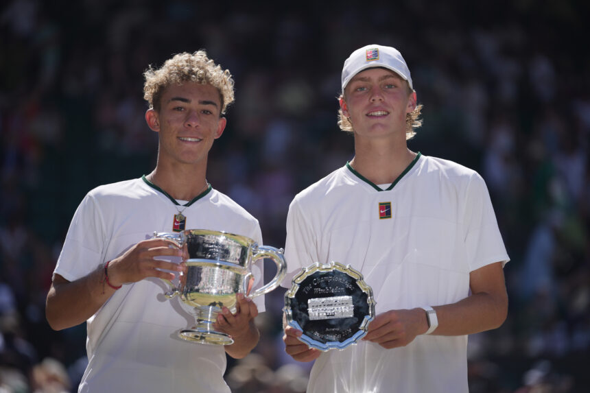 Two young athletes in white shirts pose with large trophies at a ceremony, smiling for the camera.